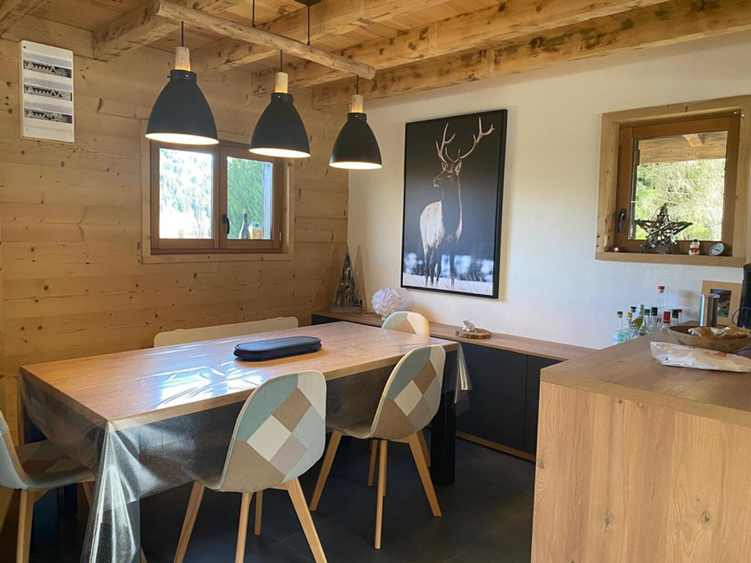 A wooden interior with a long table, chairs, pendant lights, a framed deer picture, and a window with a star decoration.