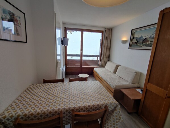 Habitación compacta con una mesa de comedor, sillas, un sofá y una mesita con una lámpara. Una ventana da a un balcón con vistas al exterior.