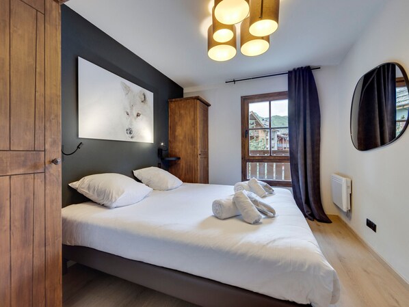 A bedroom with a bed, a wooden wardrobe, a wall-mounted artwork, a window with curtains, and a round mirror. - Ski-in Duplex with Relaxation Area (Les Arcs 1950)