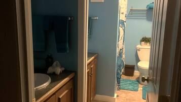 A narrow bathroom with a sink, mirror, and toilet. There is a towel rack and a small shelf above the sink. The walls are painted blue, and the floor is tiled.