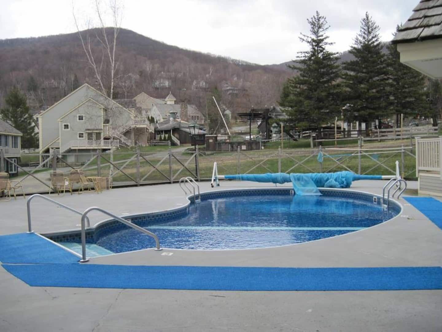 A swimming pool with a blue water slide, surrounded by a fence, with houses and a mountain in the background.