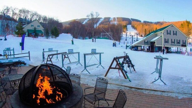 Un espacio al aire libre cubierto de nieve con un pozo de fuego, mesas y sillas.