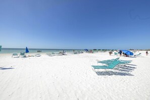 A beach with rows of blue sun loungers and umbrellas, a clear blue sky, and the ocean in the background. - Gertrude's Siesta | Bayview Unit w/ Balcony, Pools & Private Beach (Sarasota)