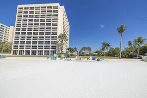 A modern hotel with a grid-like facade, a sandy beach in the foreground, and palm trees in the background. - Gertrude's Siesta | Bayview Unit w/ Balcony, Pools & Private Beach (Sarasota)