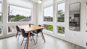 A modern dining area with a wooden table and black chairs, large windows offering views of the neighborhood, and a kitchen visible through the window. - Highline Hideaway | Corner 1BR LoHi Loft | Quiet | Dog-Friendly (Denver)