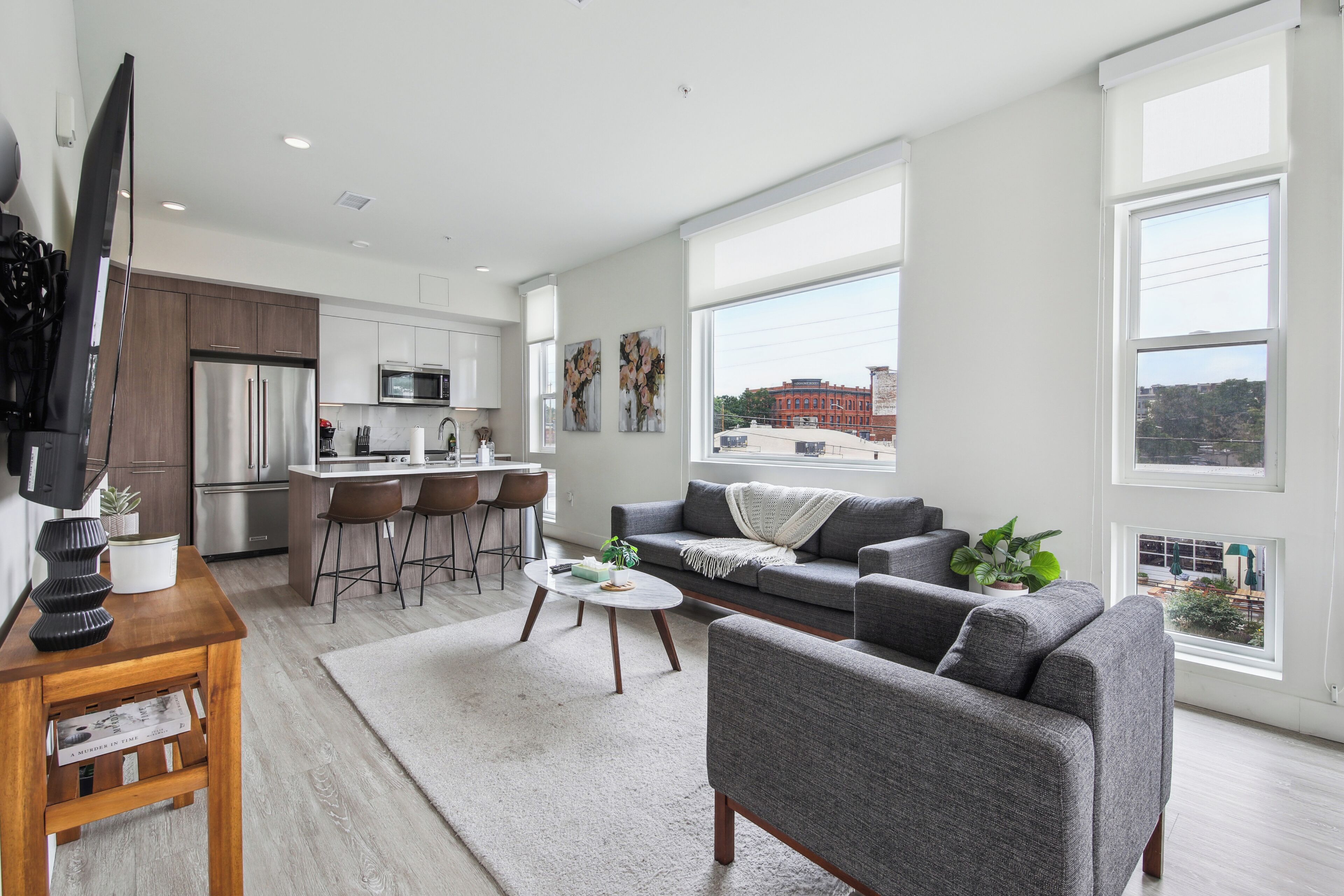 A modern living room with a grey sofa, a coffee table, and a TV on a wooden stand. There is a kitchen area with a fridge, microwave, and bar stools. Large windows offer a view of the city.