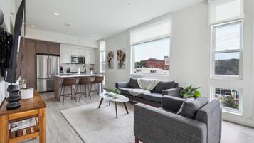 A modern living room with a grey sofa, a coffee table, and a TV on a wooden stand. There is a kitchen area with a fridge, microwave, and bar stools. Large windows offer a view of the city.
