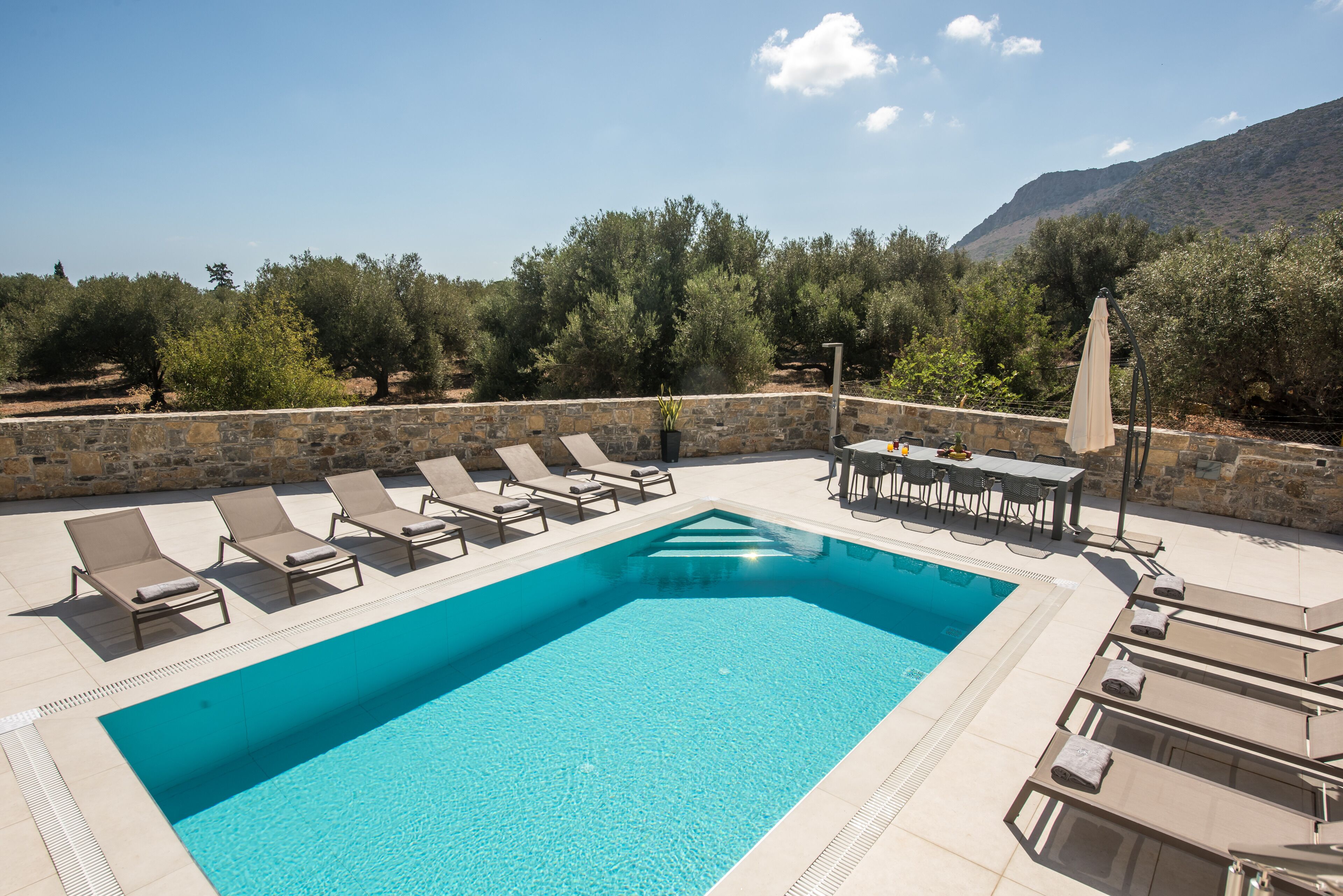 Una piscina rectangular con sillas de descanso y una mesa, rodeada de muros de piedra y olivos, con una montaña al fondo.