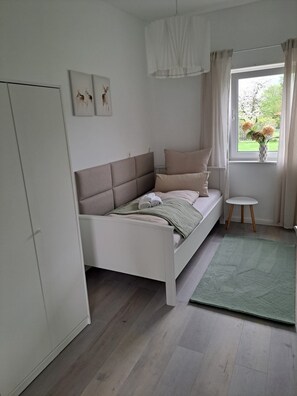 Relax in this serene bedroom with a single bed, white wardrobe, and bedside table, surrounded by natural light and a lush green rug.