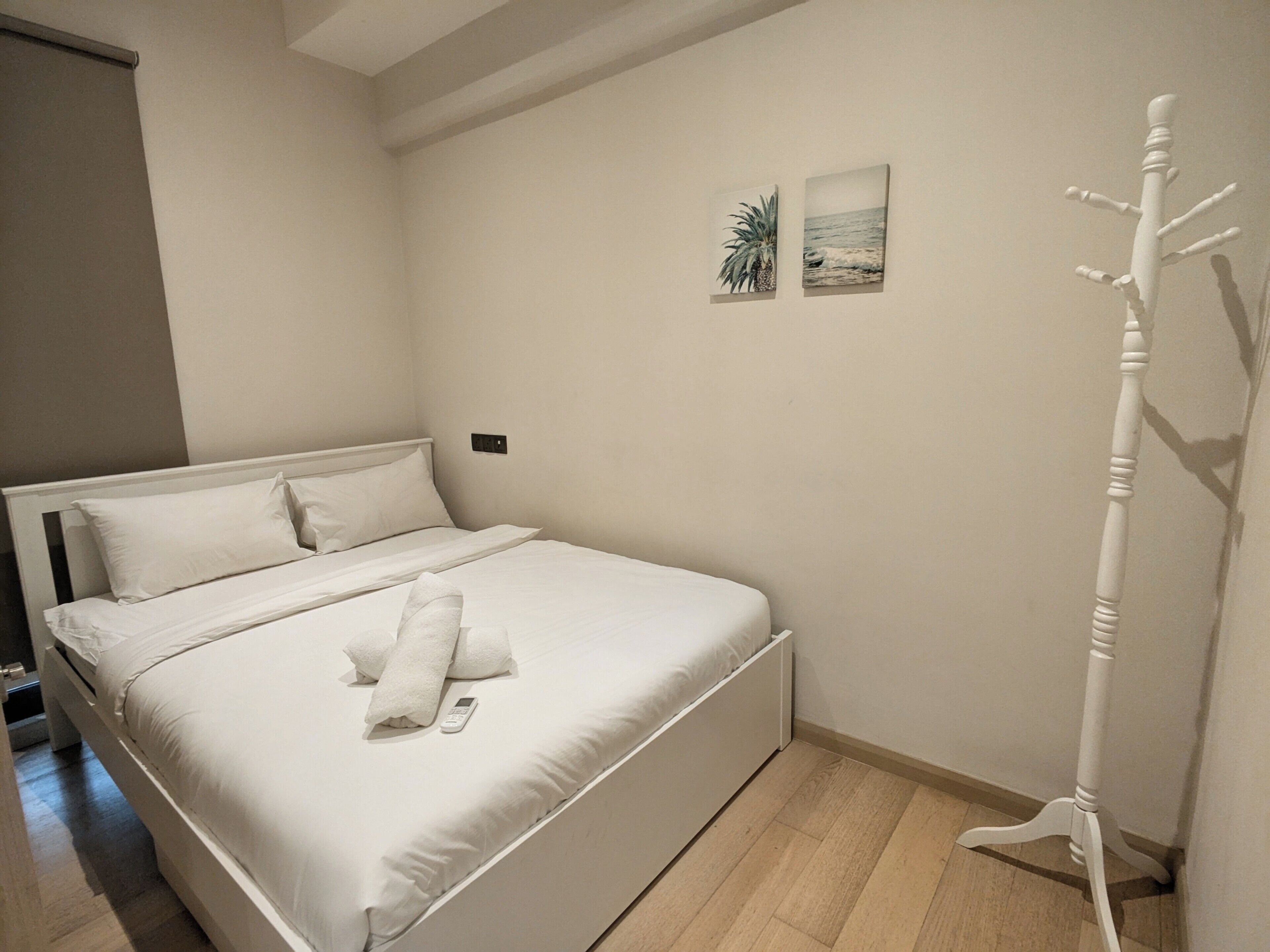 Een netjes opgemaakt bed met witte lakens, een afstandsbediening, een witte kapstok en twee kleine wanddecoraties.