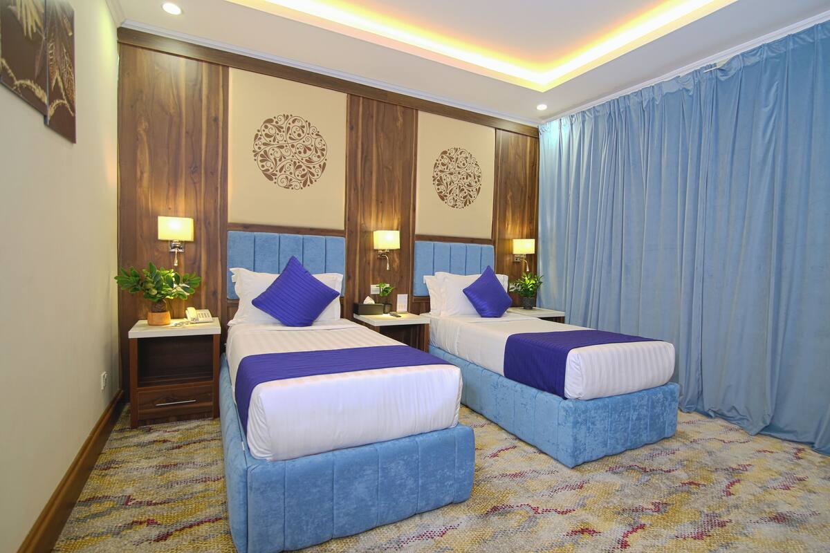 A hotel room with two beds, each with a blue headboard, white bedding, and purple pillows. There are bedside tables with lamps and a small plant on each. The room has a patterned carpet and a large window with blue curtains.