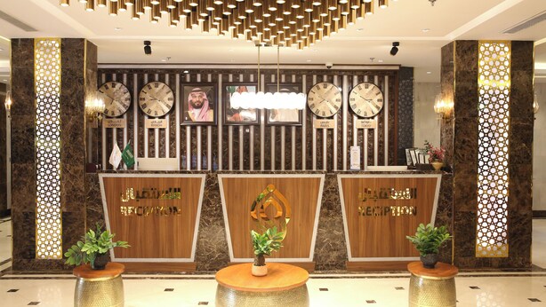 A reception area with a wooden reception desk, multiple clocks, and decorative plants. - Vandal Hotels (Jizan)