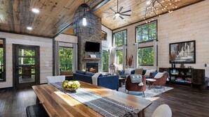 A spacious living room with a wooden ceiling, large windows, a fireplace, and a dining area with a wooden table and chairs. - Family Fun Cabin: Hot Tub + Game Room Retreat (Broken Bow)