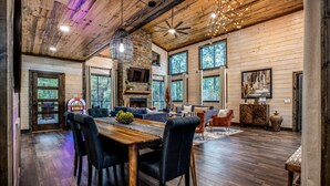 A spacious living area with a wooden dining table, multiple chairs, a fireplace, and large windows offering a view of the outdoors. - Family Fun Cabin: Hot Tub + Game Room Retreat (Broken Bow)