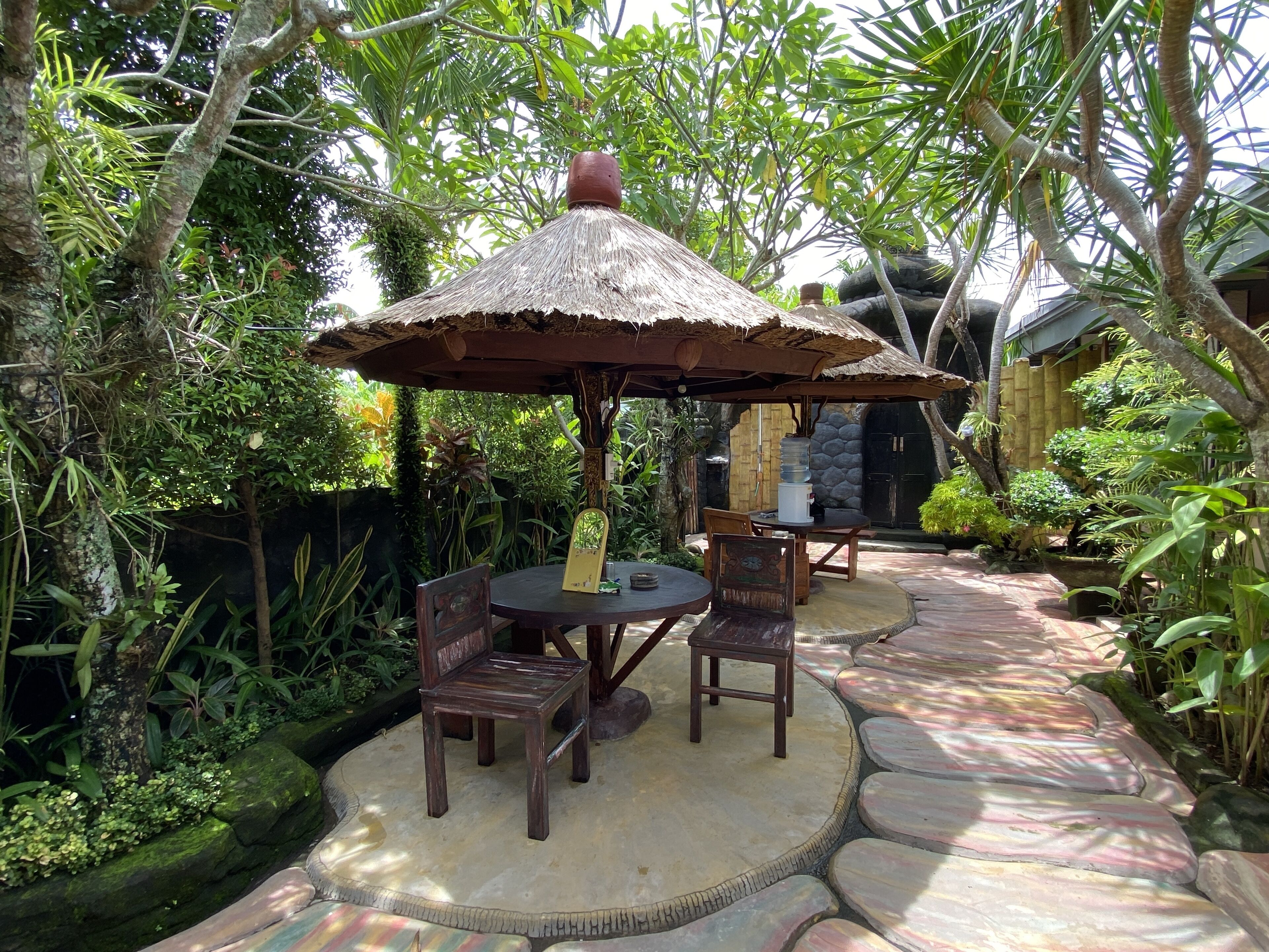 An outdoor seating area with wooden tables and chairs, surrounded by lush greenery and a thatched umbrella.
