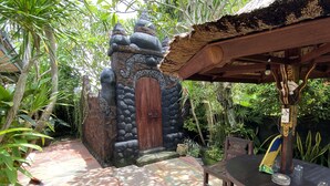 Uma área externa rústica com entrada de pedra, mesas de madeira e estrutura de telhado de palha.