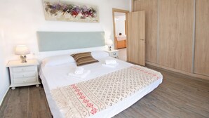 A bedroom with a bed, bedside tables, a nightstand, a lamp, a hanging light fixture, a wardrobe, and a painting on the wall.