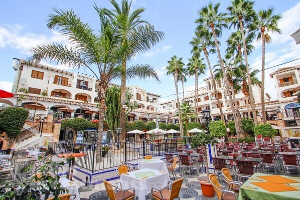 An outdoor café area with tables and chairs, surrounded by palm trees and buildings with balconies.