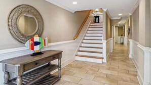 A hallway with a staircase, a round mirror, and a wooden console table. - The Tip of Tybee Townhouse (Tybee Island)