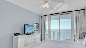 Une chambre avec un grand lit, un téléviseur posé sur une commode, un balcon avec vue sur la mer et un ventilateur de plafond.