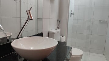 Egy modern fürőszoba, melyben egy nagy, fehér mosdókagyló, egy fekete mosdópult, egy fehér WC és egy zuhanyzó található.