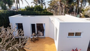 A modern white house with a wooden deck, a patio with a table and chairs, and a small garden area with plants.