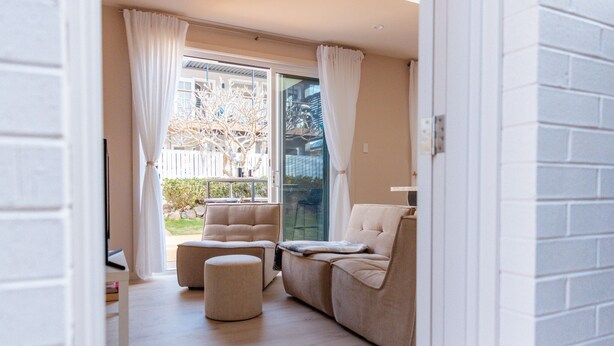 A modern living room with a beige sofa, a round ottoman, and a glass door leading to a balcony.