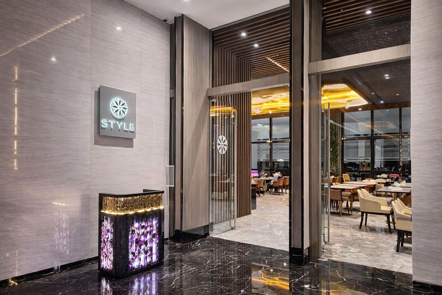 A modern hotel lobby with a reception desk, a sign reading 'STYLE', and a dining area with tables and chairs.