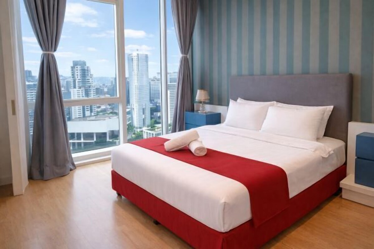 A hotel room with a large bed, a nightstand, a lamp, and a view of the cityscape through large windows.