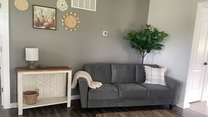 A living room with a grey sofa, a wooden side table, a woven rug, and a green plant. - The Blue Door Place
 (Florence)