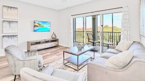 A modern living room with a sofa, armchairs, a coffee table, and a TV. There is a balcony with a view of trees and a railing. - Upstay - LRG Condo w Outdoor Pool, Sauna (Winter Garden)