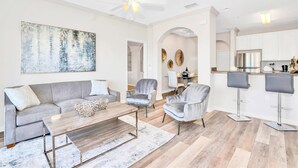 A modern living room with a grey sofa, a coffee table, and two armchairs. There is a kitchen area with white cabinets and a bar stool. - Upstay - LRG Condo w Outdoor Pool, Sauna (Winter Garden)