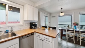 Una cocina moderna con armarios blancos, encimera de madera y lavavajillas. Hay un comedor con una mesa y sillas, y vistas al océano a través de grandes ventanales.
