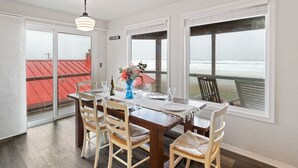 Un comedor con una mesa de madera, cuatro sillas, un jarrón con flores y vistas al océano a través de amplias ventanas.