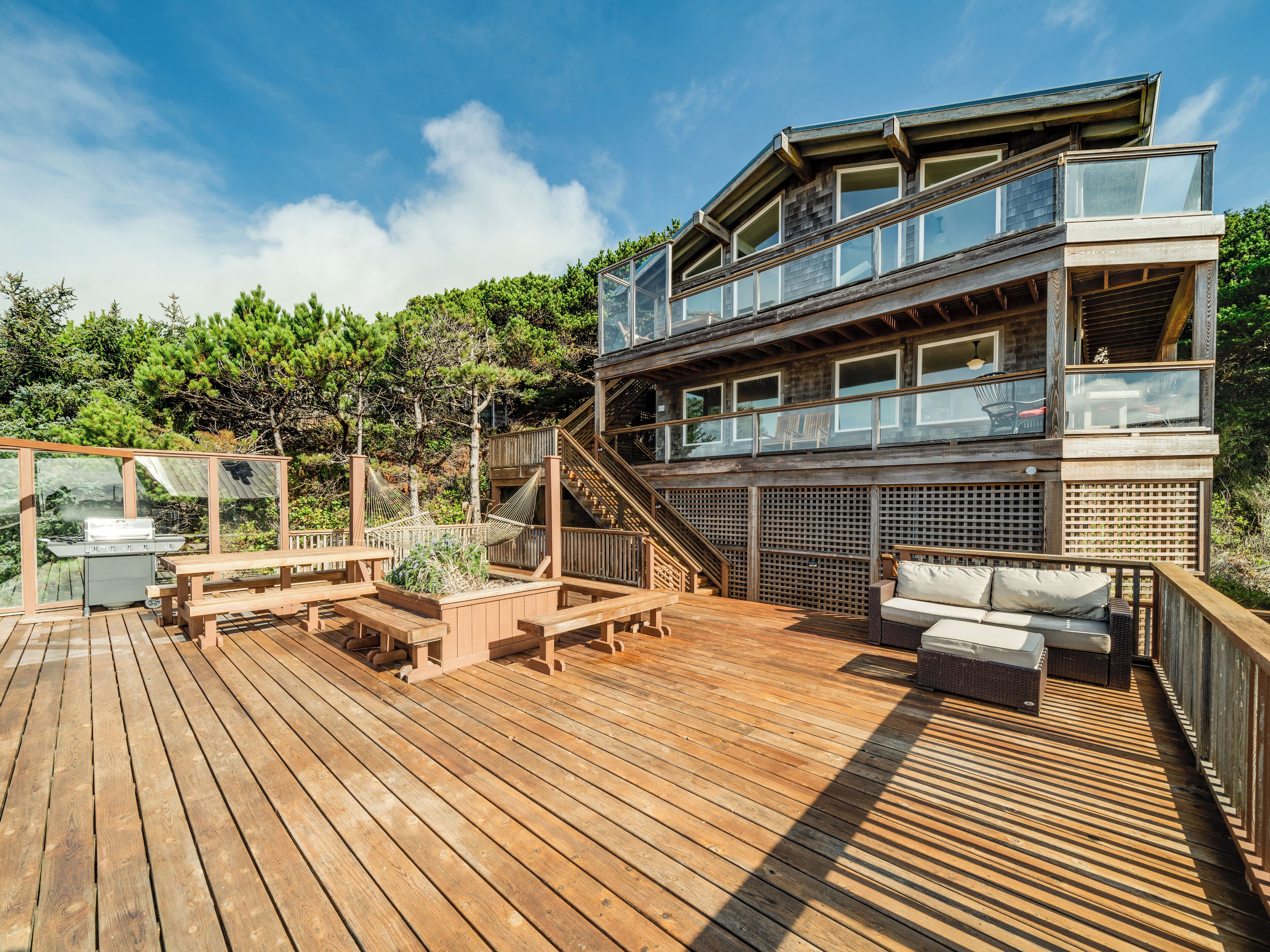 Una terraza de madera con mobiliario exterior, una parrilla y una escalera que lleva a un balcón con barandillas de vidrio.