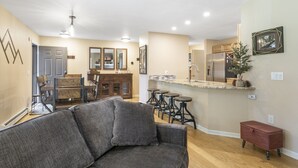 A modern living room with a gray sofa, a dining area with wooden chairs, a kitchen with a refrigerator, and a small red cabinet. - 901 Crazy Horse Circle 2 Bedroom Condo by RedAwning (Edwards)