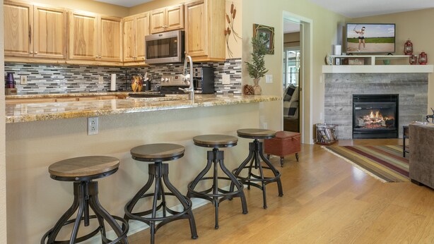 A modern kitchen with wooden cabinets, a microwave, and a granite countertop. There are three stools and a fireplace in the living area. - 901 Crazy Horse Circle 2 Bedroom Condo by RedAwning (Edwards)