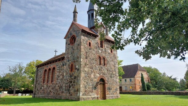 Una pequeña iglesia de piedra con una torre de campanas, ventanas de arco y una cruz en la parte superior.