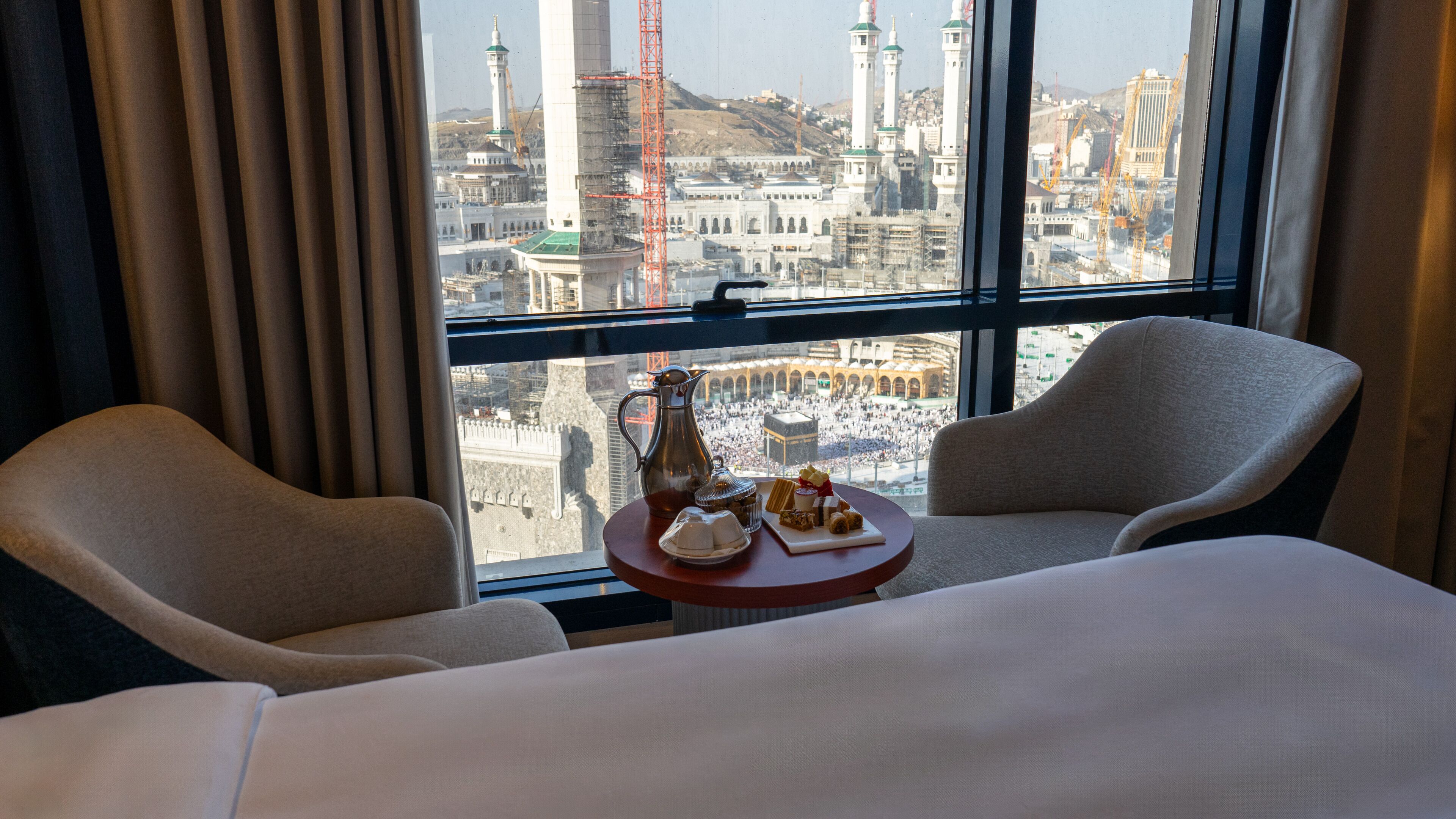 Une chambre d’hôtel avec vue sur le paysage urbain, comprenant une mosquée et des grues de construction. On y trouve deux chaises et une petite table avec une carafe et des en-cas.
