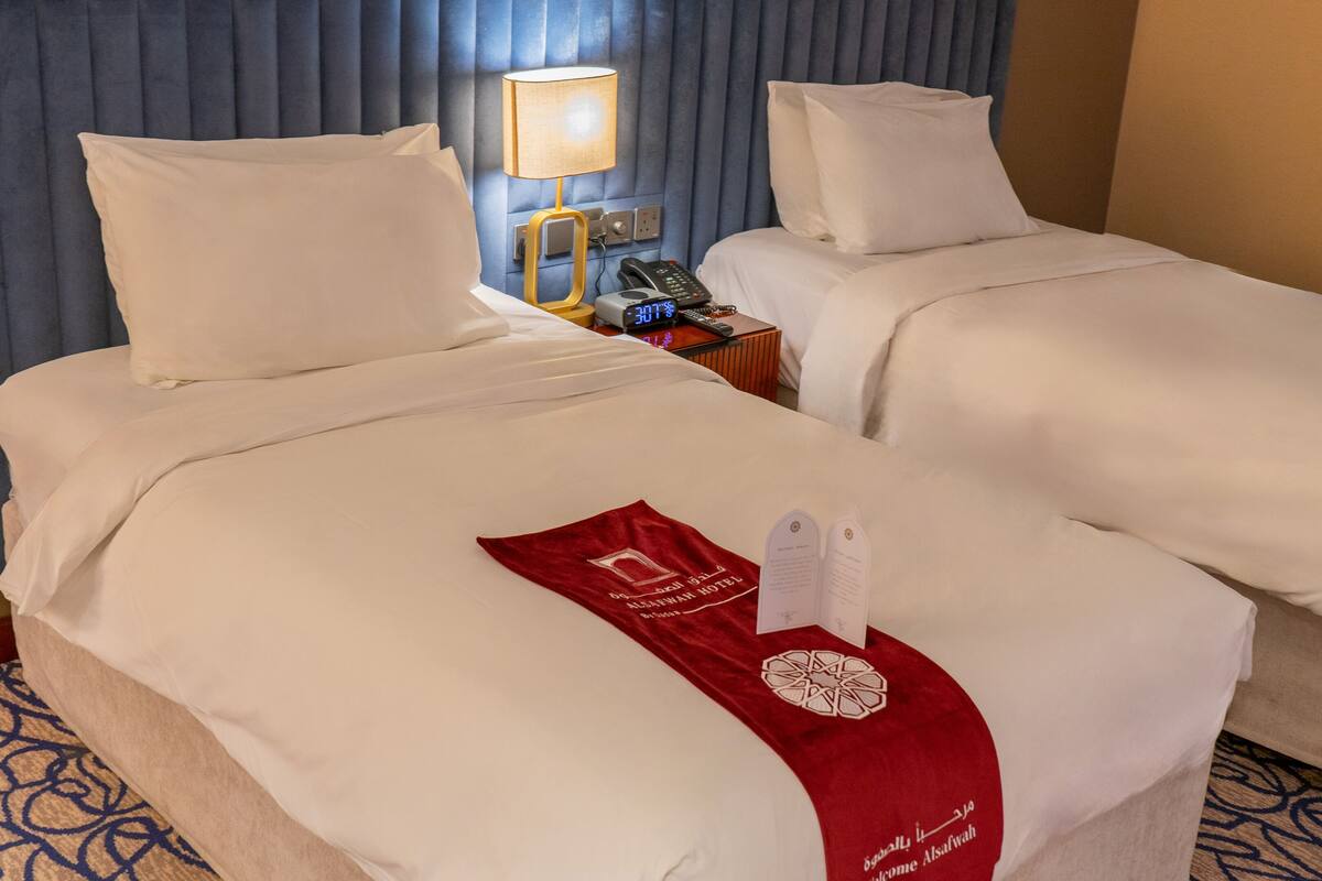 A hotel room with two beds, a nightstand with a lamp, a phone, and a digital clock.