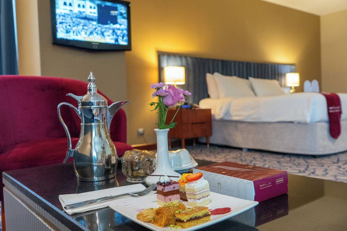A hotel room with a bed, a red armchair, a coffee table with a plate of food, a vase with flowers, and a television on the wall.