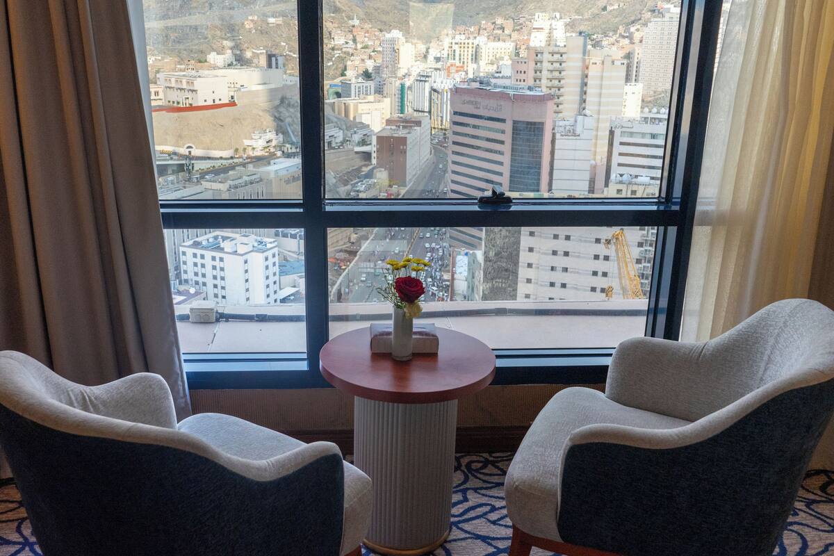 A hotel room with two armchairs, a small table with a vase of flowers, and a large window offering a city view.