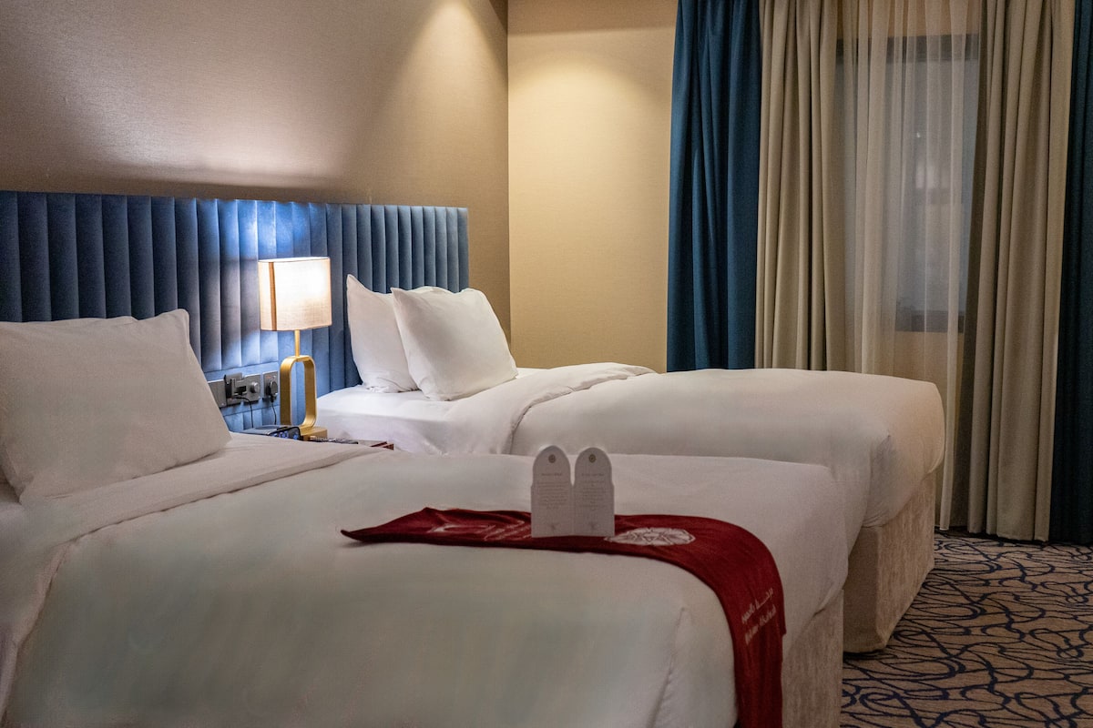 A hotel room with two beds, a blue headboard, a red throw blanket, and a patterned carpet.