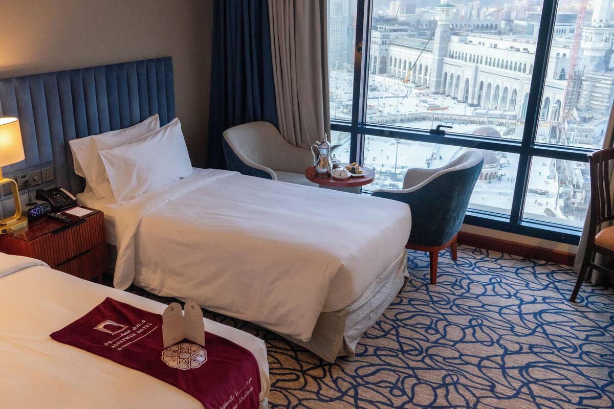 A hotel room with two beds, a nightstand with a lamp, a chair, and a view of a cityscape through large windows.