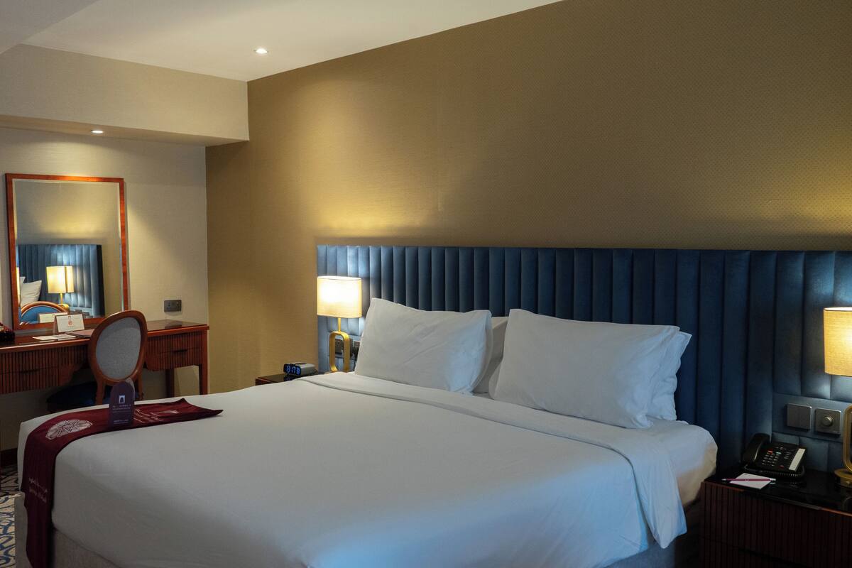A hotel room with a large bed, a desk with a mirror, two bedside lamps, and a nightstand with a phone and a lamp.