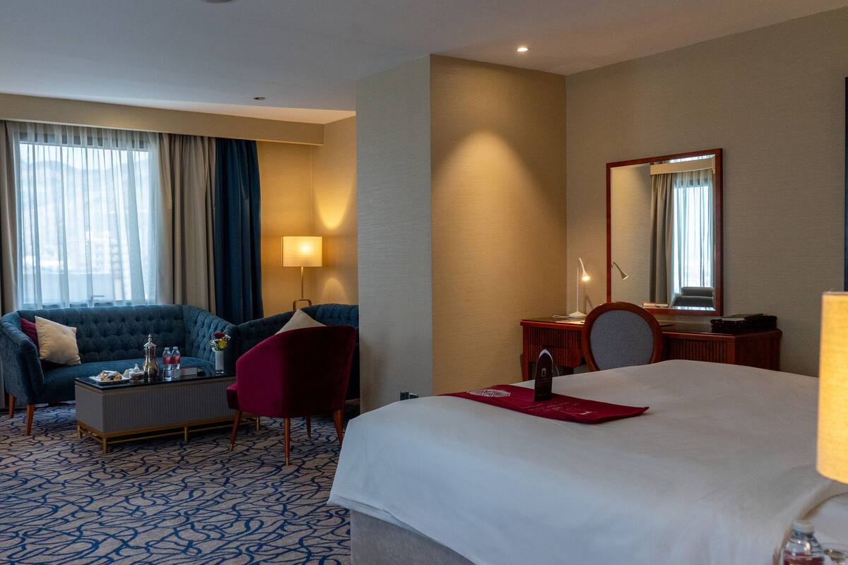 A hotel room with a large bed, a sofa, a desk, and a chair. There is a window with curtains, a lamp, and a mirror on the wall.