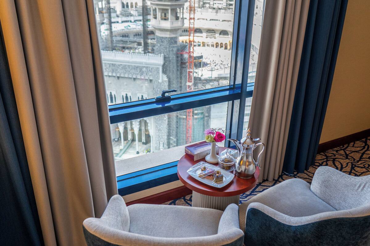 A hotel room with two armchairs, a small table with a vase of flowers, a tray of pastries, and a view of a cityscape through a large window.