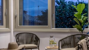 Une pièce avec deux fauteuils en osier, une petite table sur laquelle se trouvent une carafe et des verres, et une grande fenêtre donnant sur des arbres et un ciel dégagé.