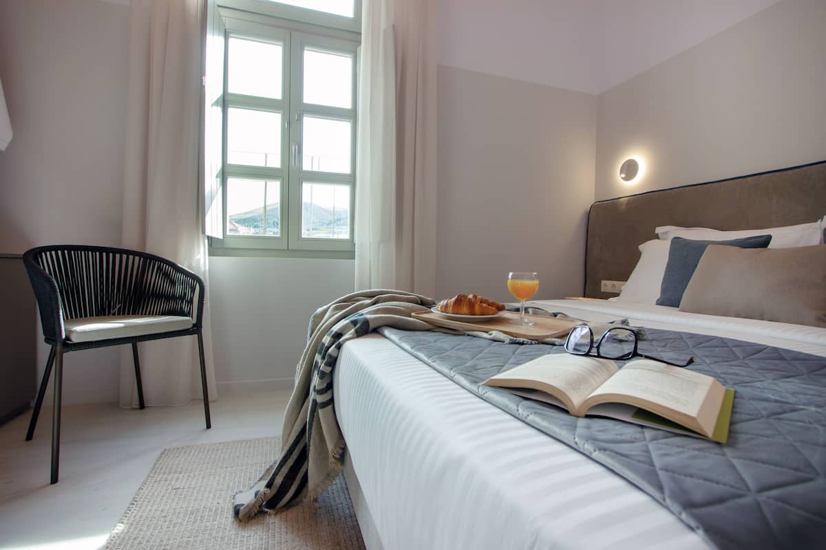 Habitación de hotel ordenada con cama, una silla, una ventana con cortinas y una bandeja de desayuno con un croissant y jugo de naranja.