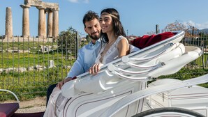Un couple est assis dans une voiture ancienne, avec en arrière-plan des ruines antiques.